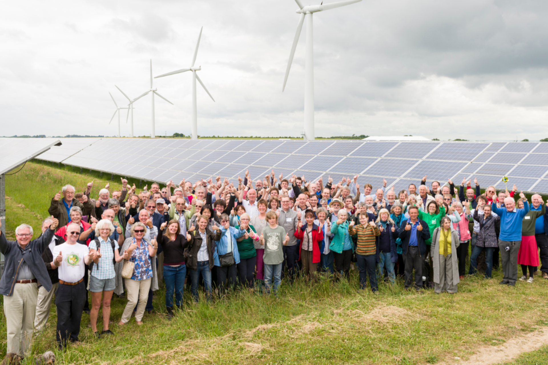 A group of people stand in front of a wind farm, with turbines in the background.