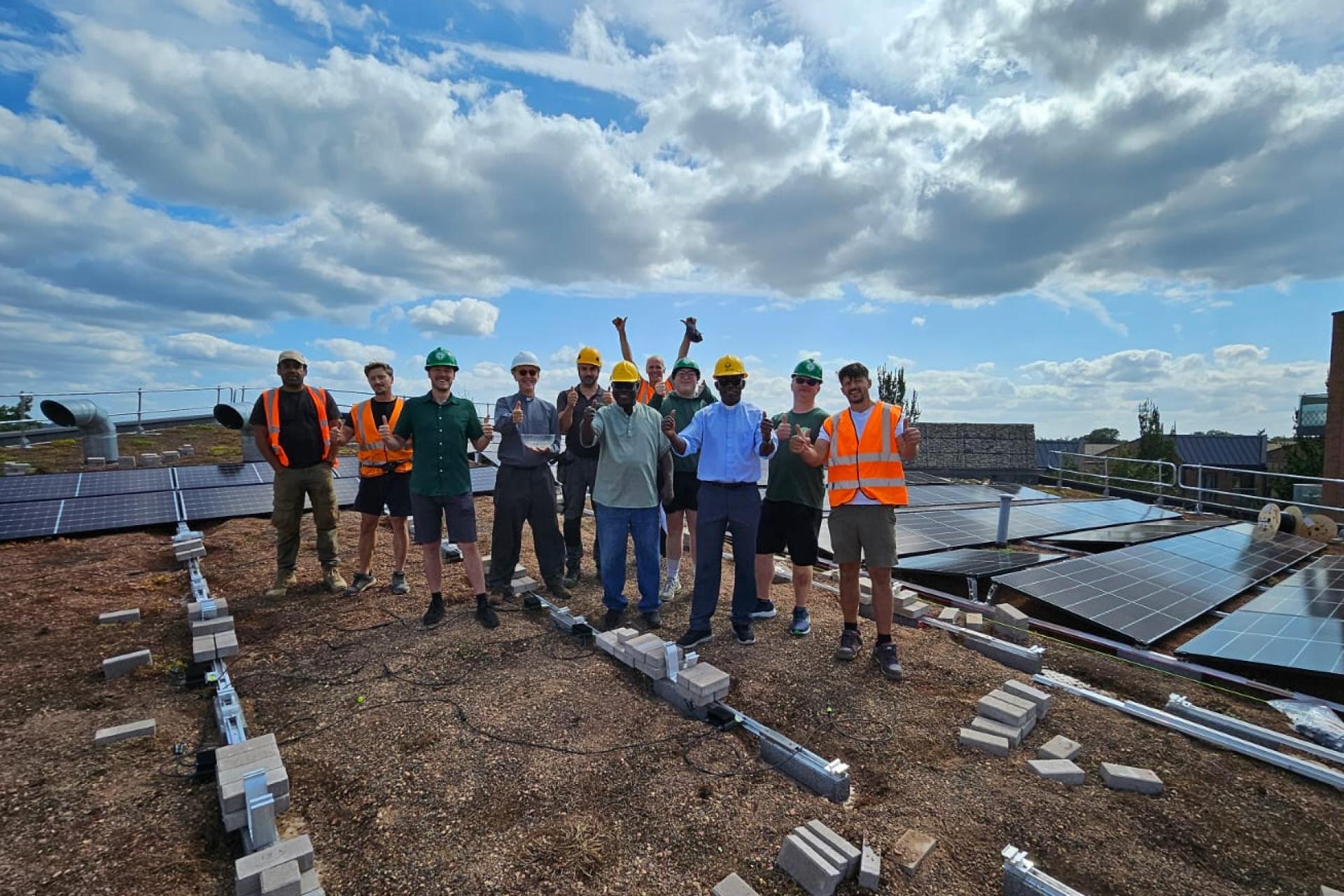 Workers in high visibility jackets and hardhats smiling on a roof alongside solar panels. 