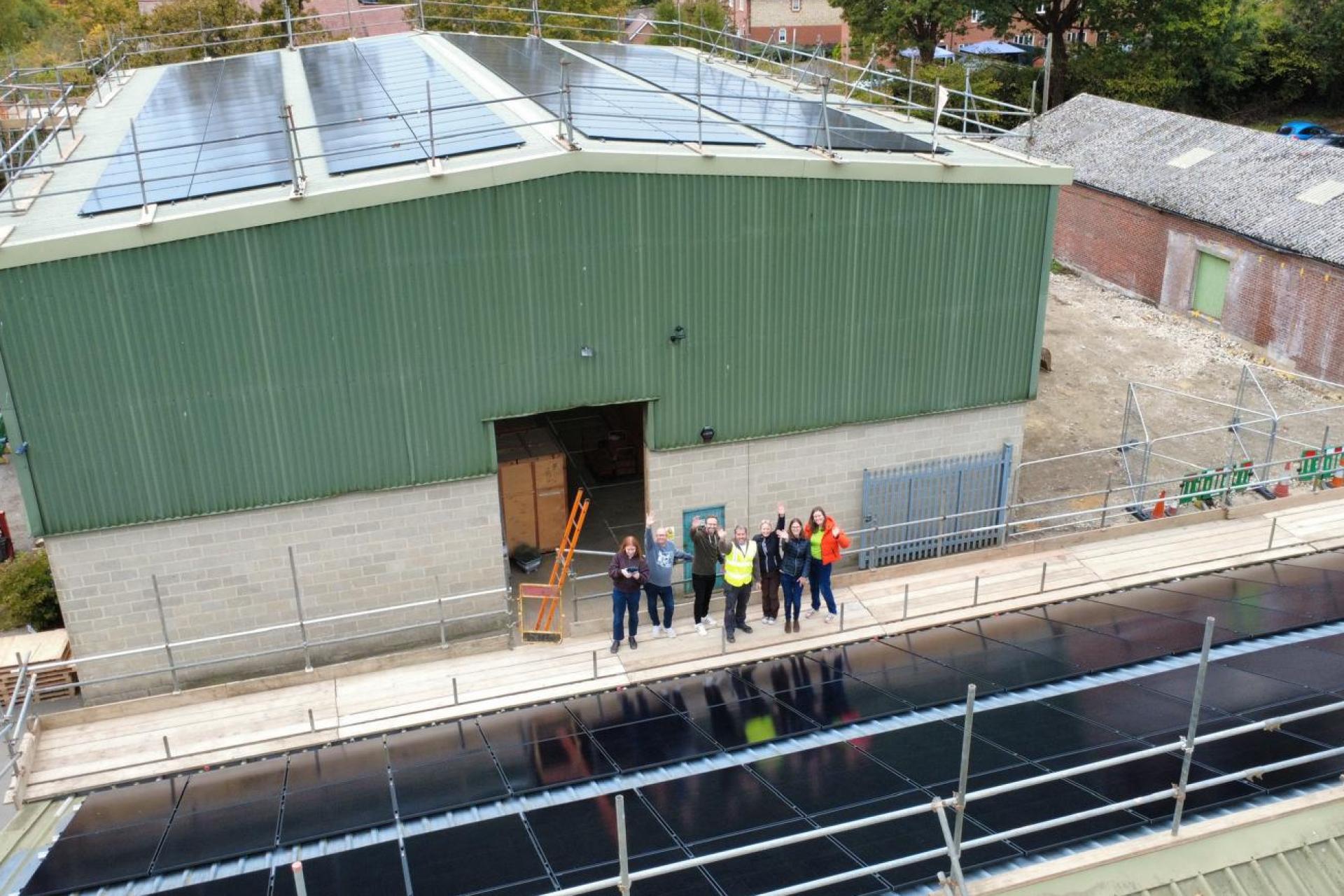 A large green warehouse with solar panels on the roof at 6 members of staff stood proudly in front of the building.
