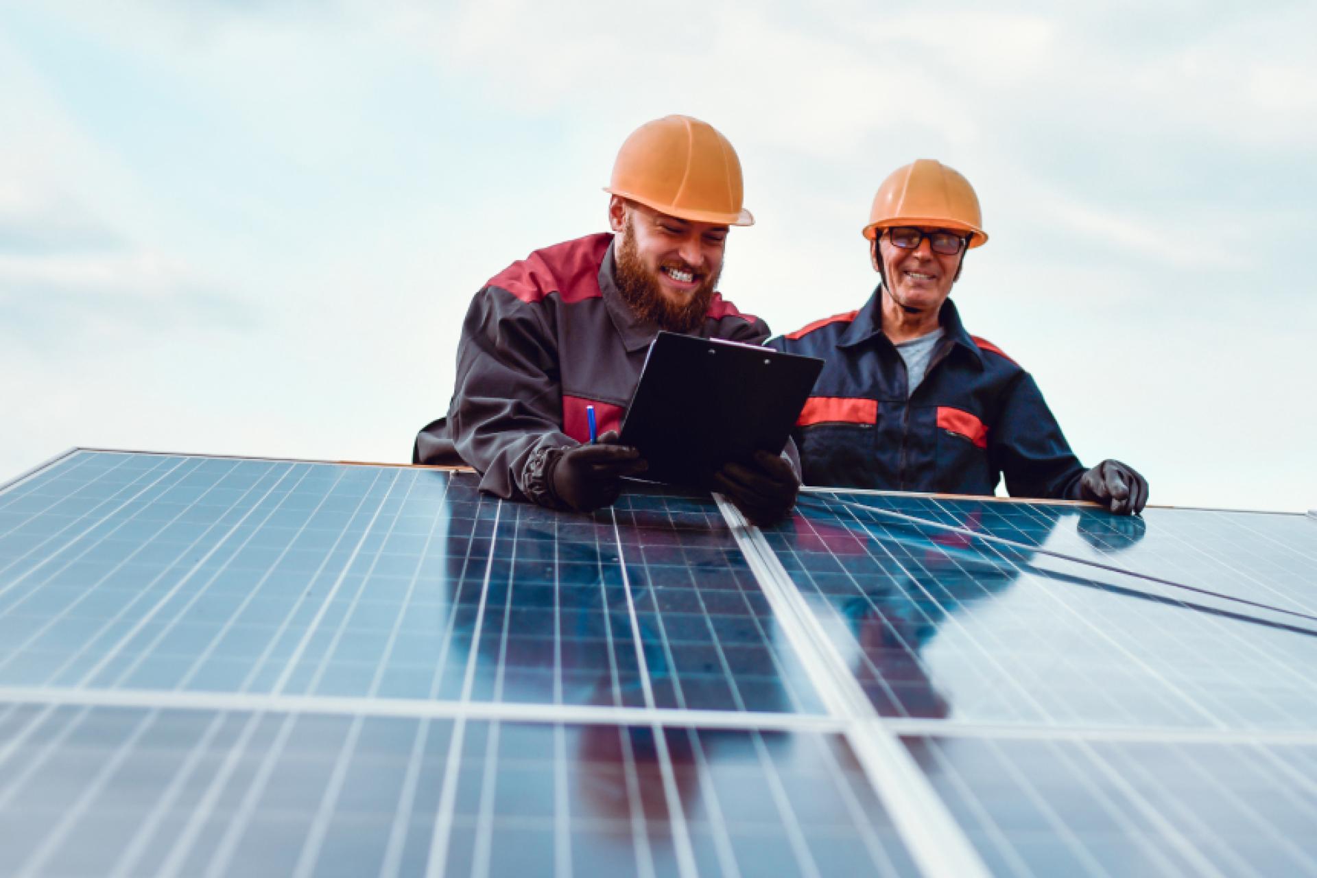 Two workers wearing hard hats look down at solar panels and smile.